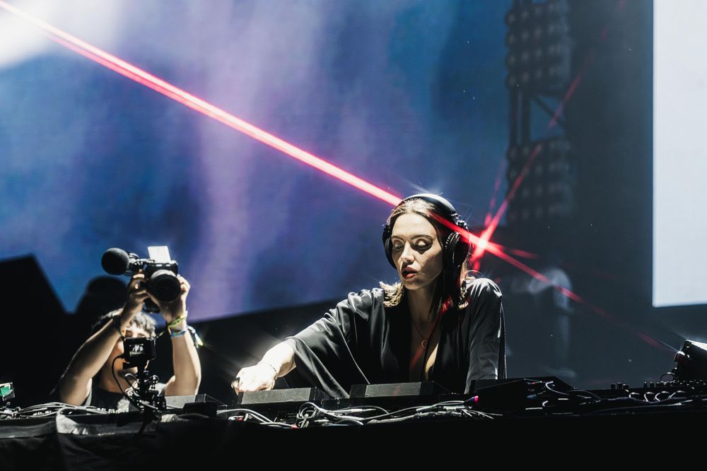 DJ performs at a dark concert venue with dramatic red lighting and electronic equipment.