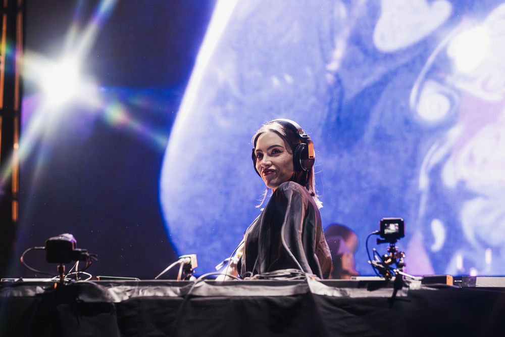DJ performing at an electronic music event with blue stage lighting and visual effects in the background.
