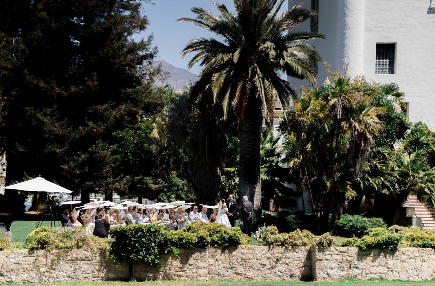 A panoramic view of a lush garden with palm trees, stone walls, and white umbrellas in a Mediterranean setting.