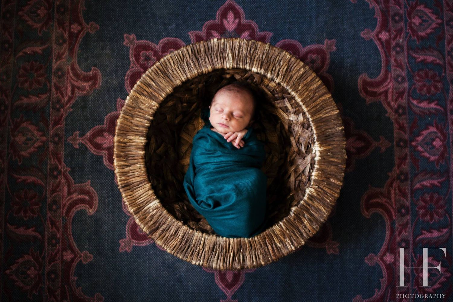 Newborn photography featuring baby sleeping in woven basket against patterned dark background.