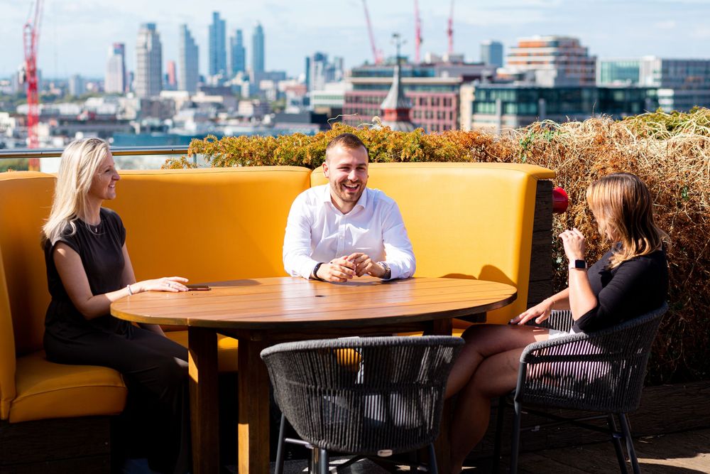People sit at tables in a restaurant with large windows overlooking a cityscape in the background.