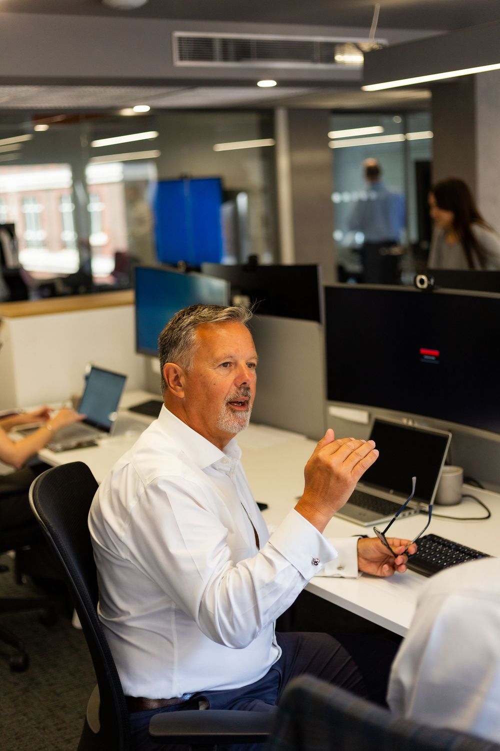 Professional in a white dress shirt gestures while speaking at a desk in a modern office space with computers.