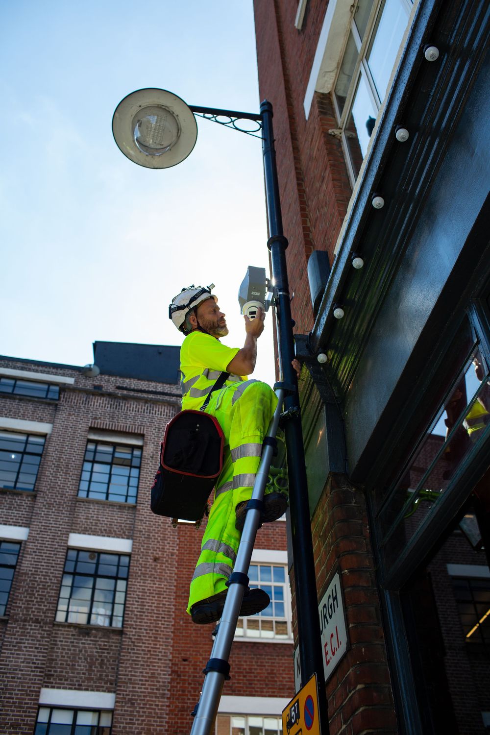 Maintenance worker in high-visibility safety gear works on a street lamp while standing on a ladder against a brick building.