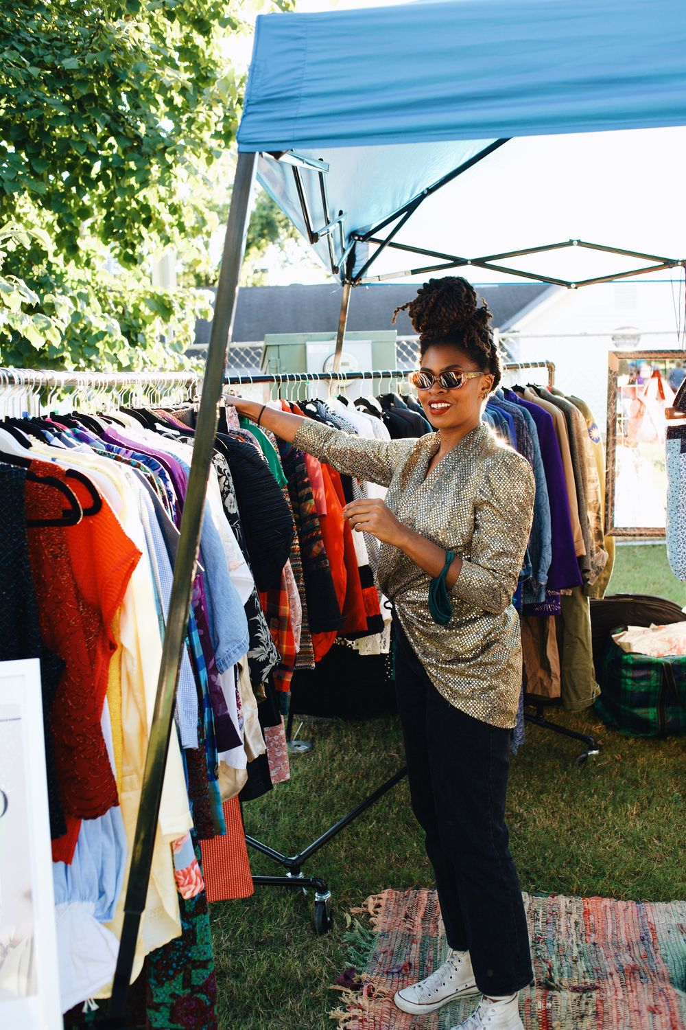 A person browses through clothing racks at an outdoor market stall under a blue canopy tent.