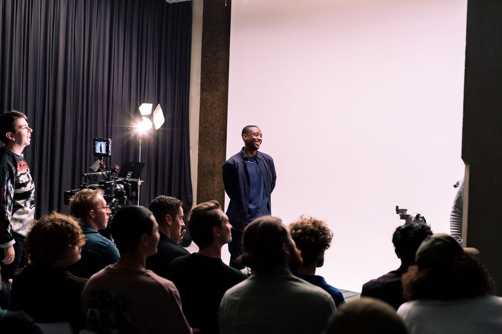 A group of people gather in a dark theater space with black curtains and stage lighting visible in the background.