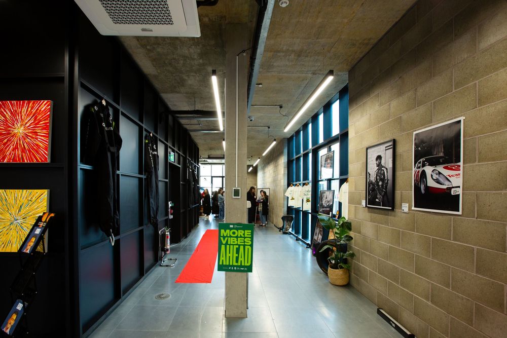 Modern office hallway with red carpet, brick walls, framed car photos, and a neon sign reading More Vibes Ahead.