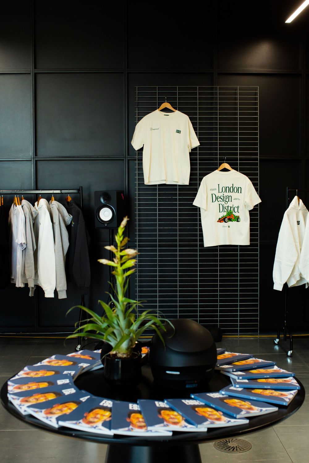 Modern retail store with London Design District t-shirts displayed on dark grid wall, clothing rack, and floral centerpiece table.