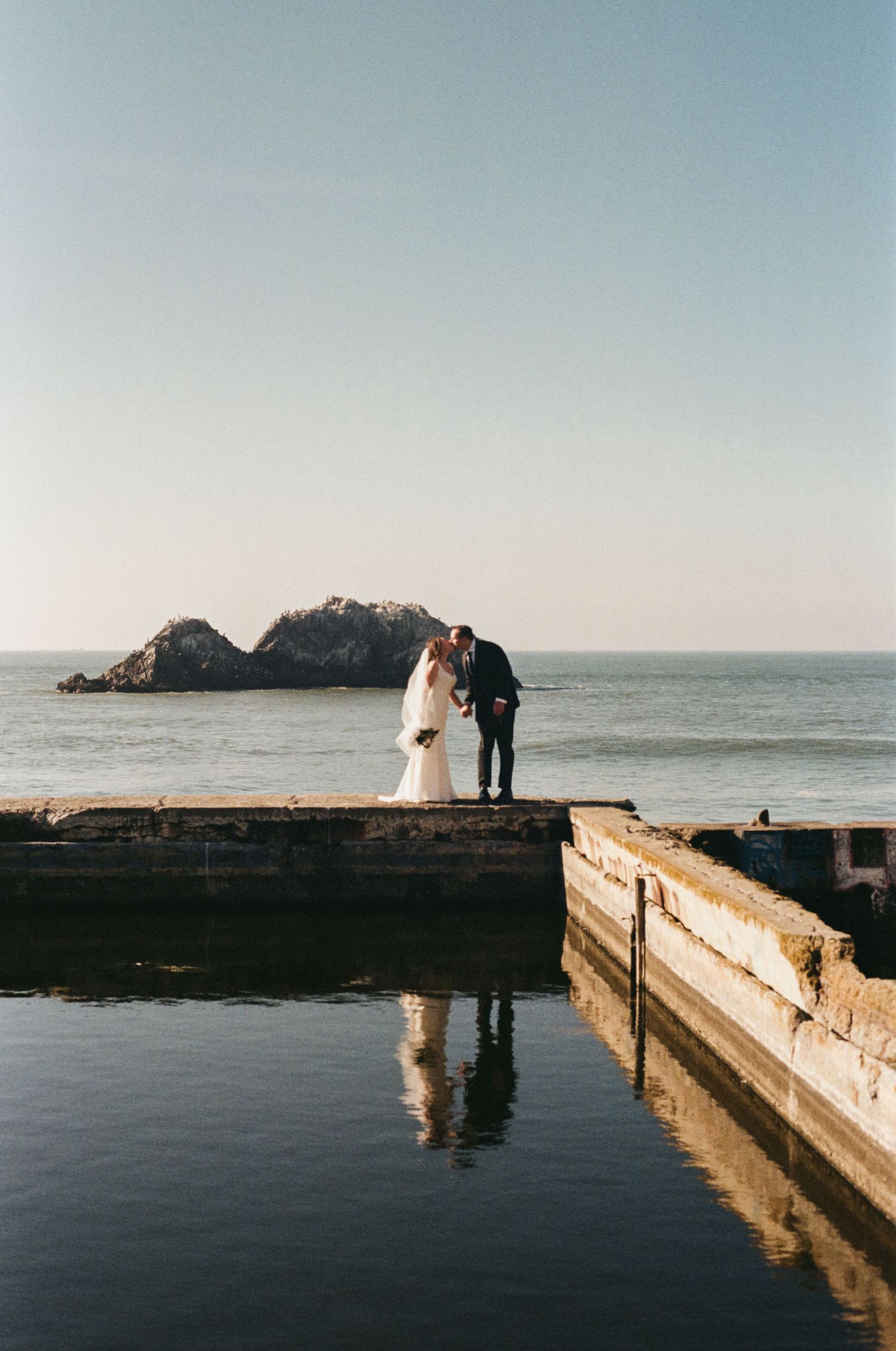 Couple stands on rocky coastal outcrop at Sutro Baths with ocean and cliffs in background during wedding photoshoot.