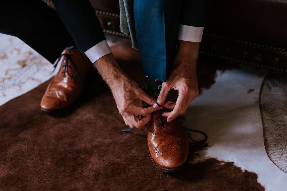 Close-up shot of brown leather dress shoes being tied with laces on wooden floor.