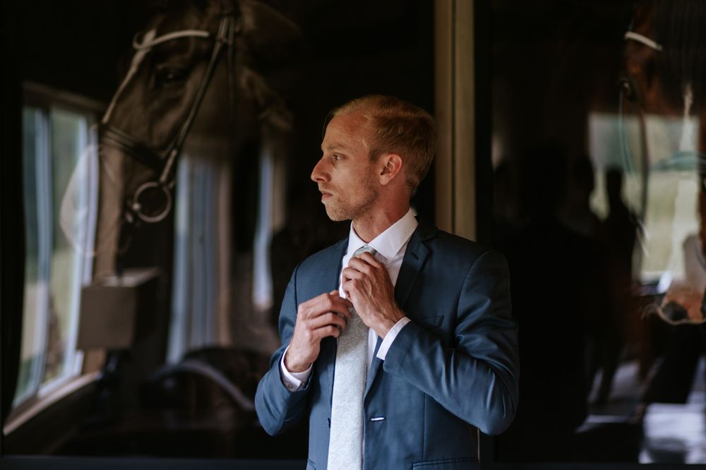 Man in navy suit adjusts tie while standing near window in moody lighting.