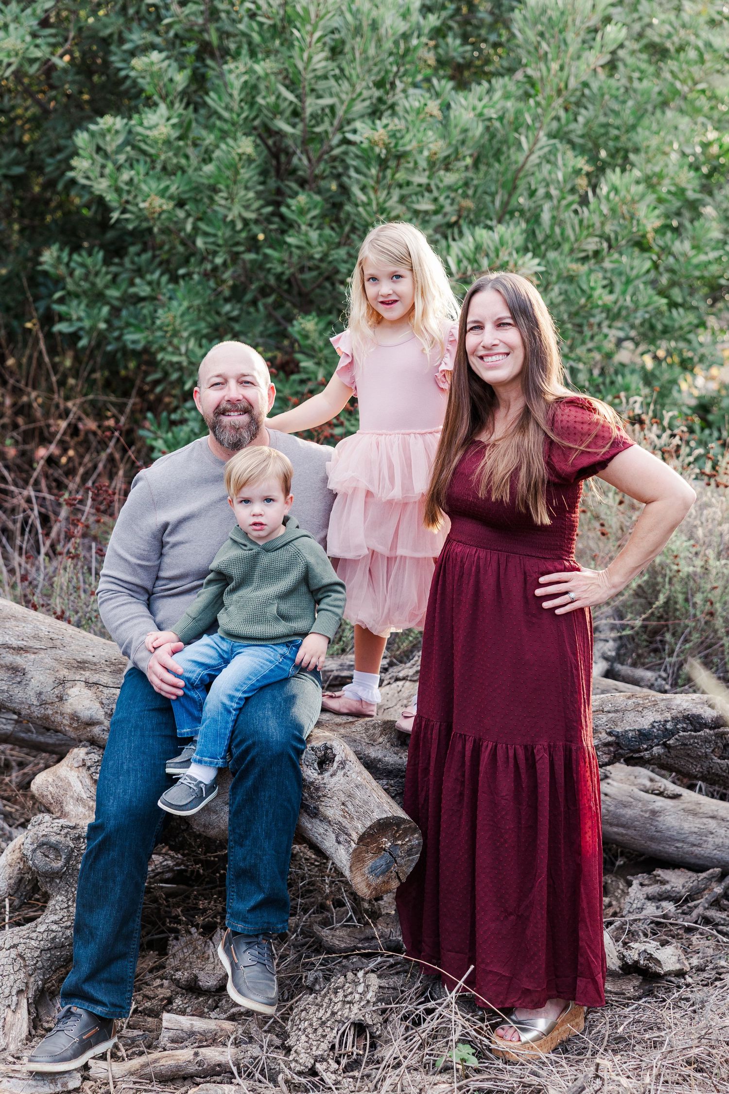 A family poses together on fallen logs in an outdoor setting with green foliage in the background.