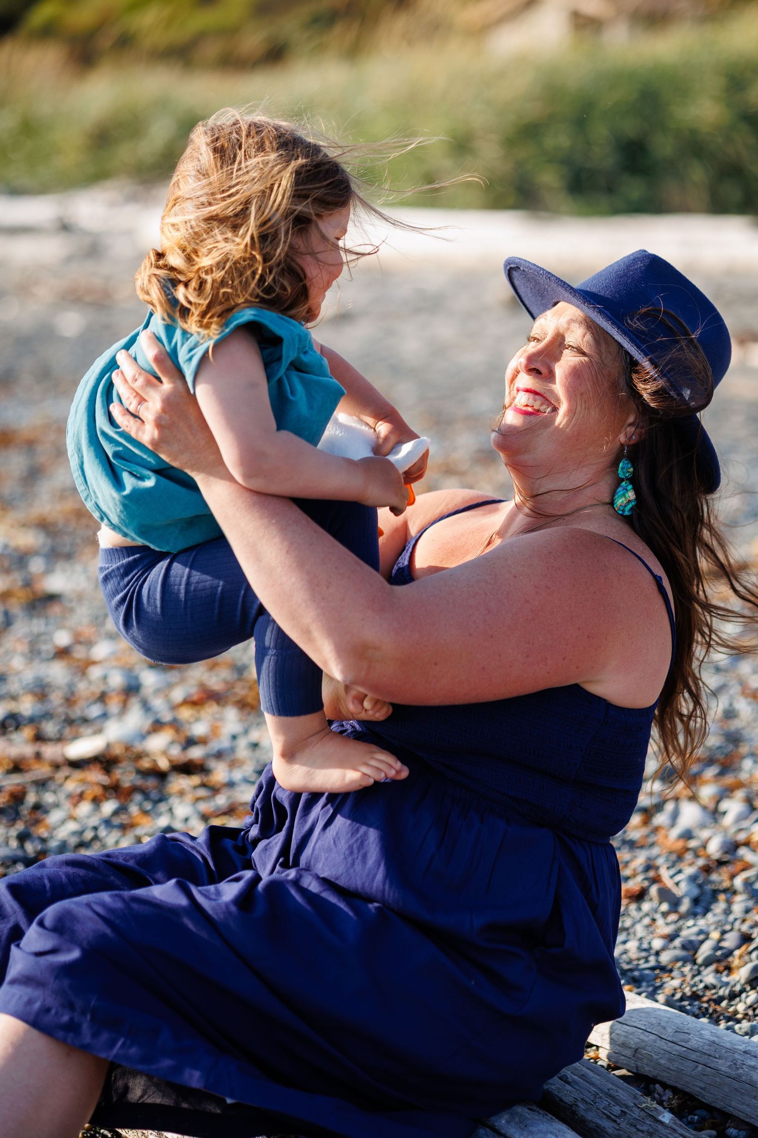 Joyful mother lifting laughing child on rocky beach, both wearing blue, sharing a playful moment.