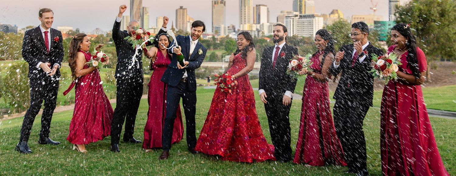 Wedding party in red and black formal attire celebrating outdoors with confetti against city skyline.