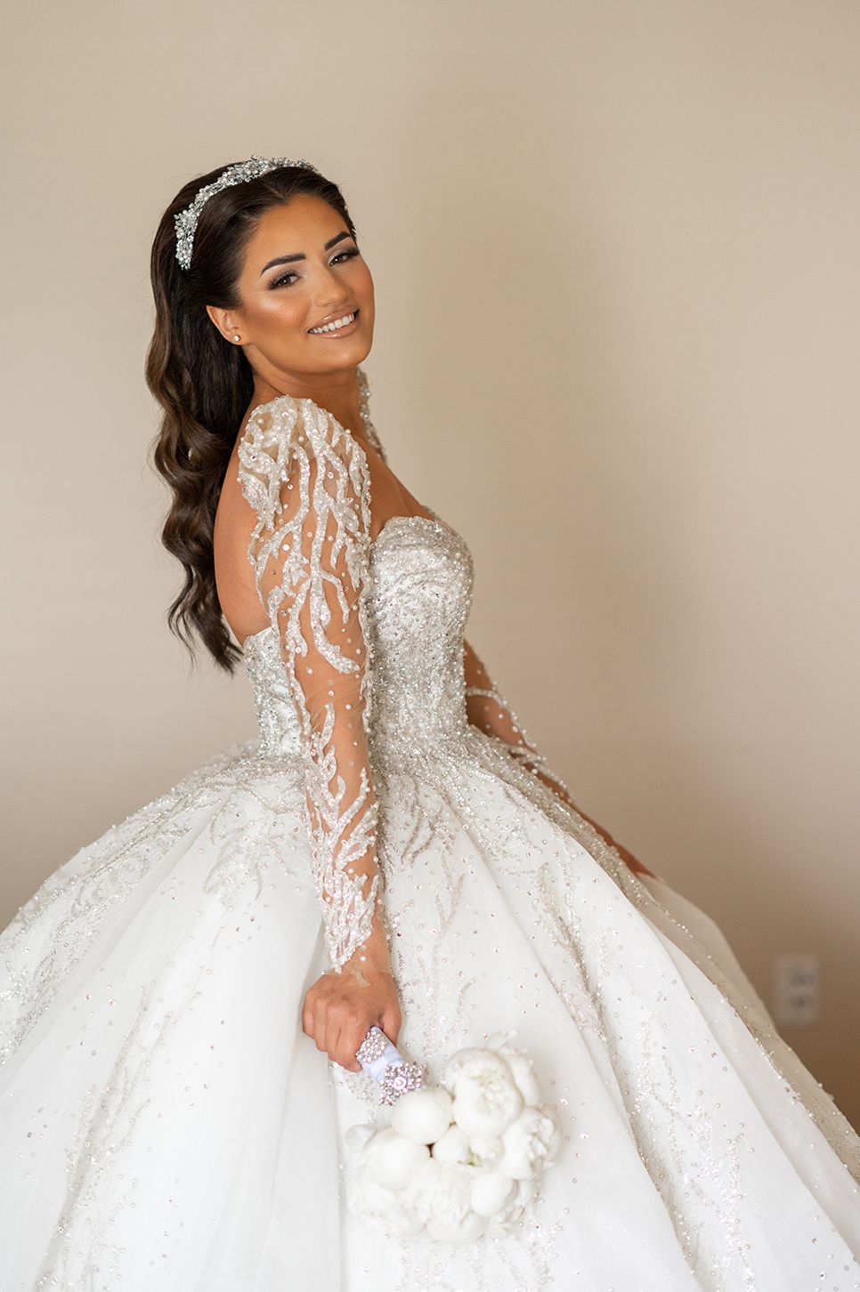 A stunning ball gown wedding dress with intricate lace sleeves and a sparkling tiara on a beige background.