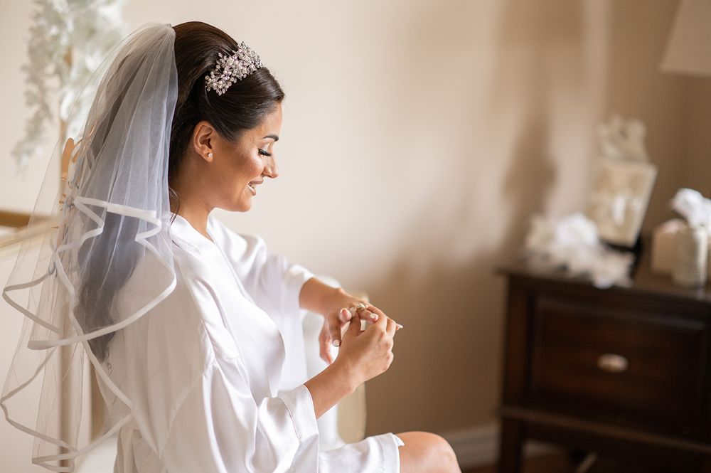 A bride in a white robe sits by a window holding a coffee cup during wedding day preparations.
