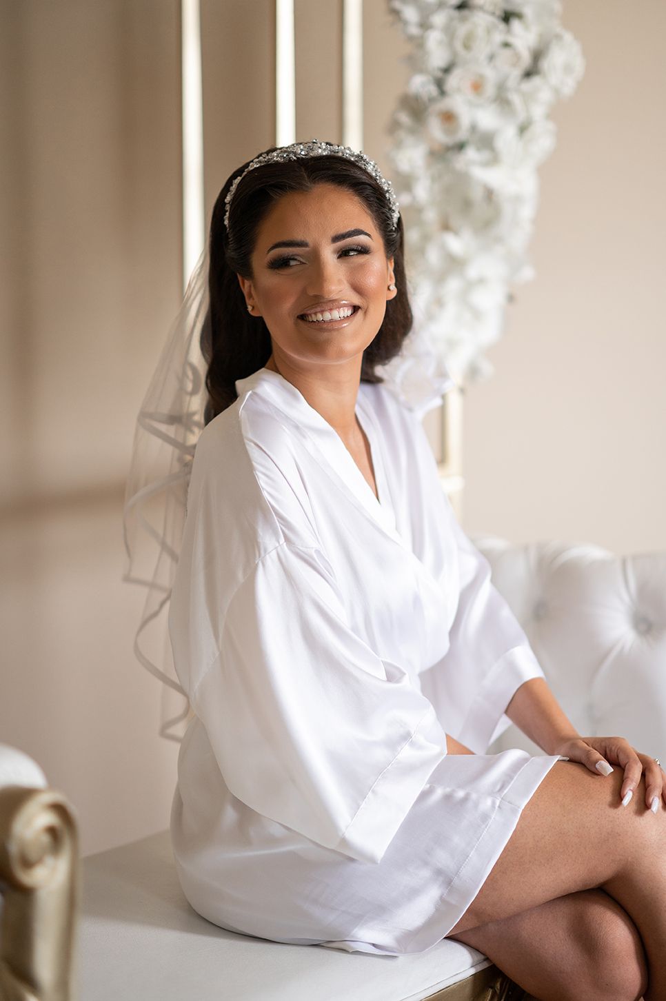 Wedding preparation scene showing someone in a white silk robe and delicate tiara sitting elegantly on a light-colored chair.