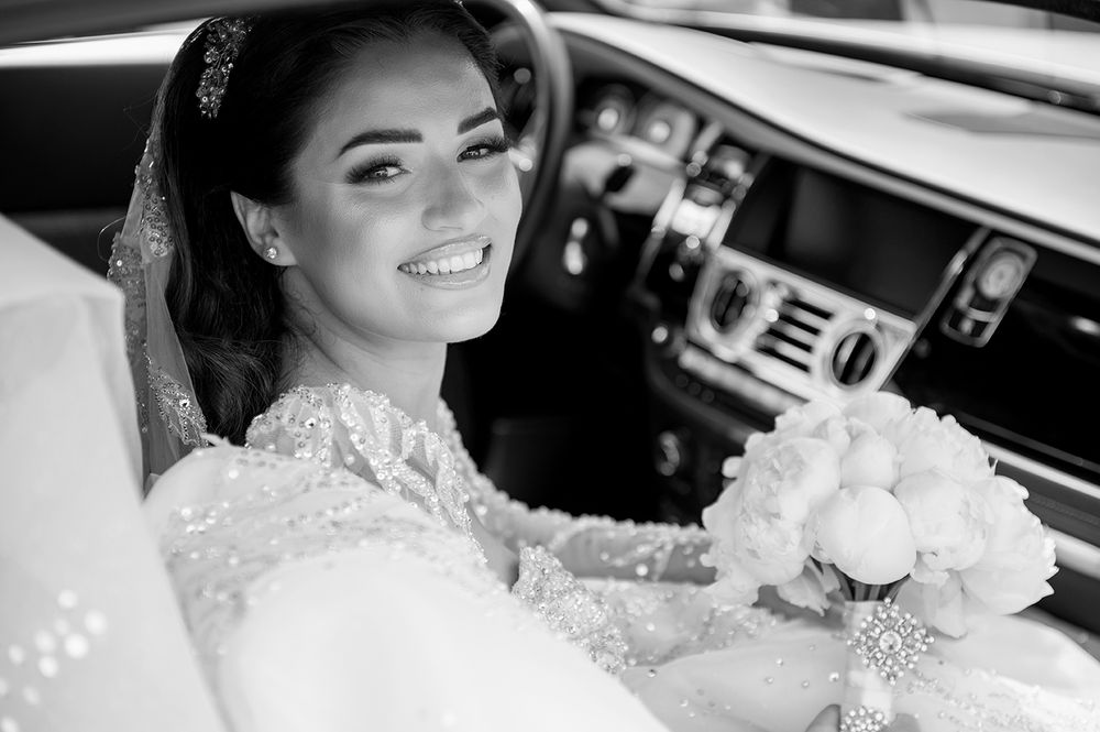 An elegant bride in a luxurious white lace gown poses in a car with a white rose bouquet.