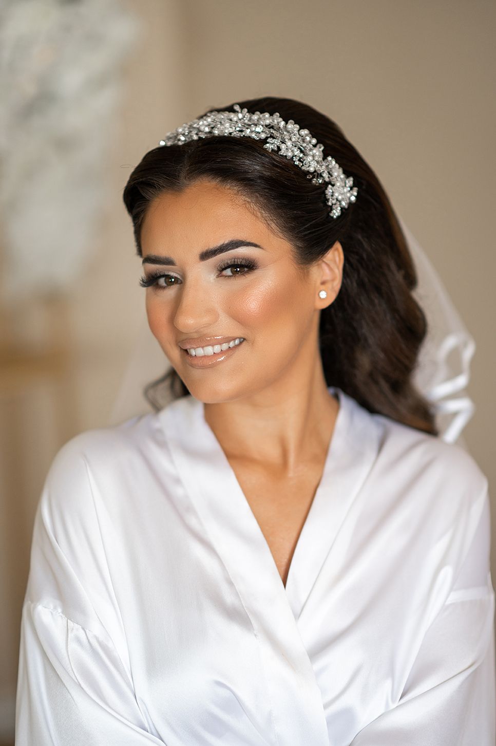 A bride wearing a white robe and sparkling hair accessory smiles while getting ready for her wedding day.