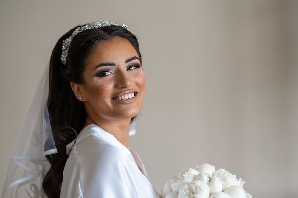 A bride in a white robe smiles while holding a white bouquet, wearing a crystal headpiece and veil.