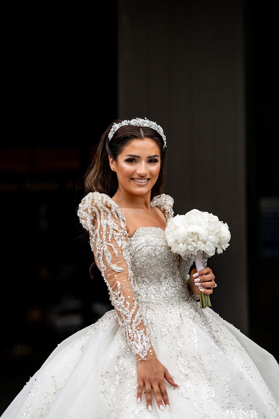 A bride in a luxurious white lace gown with sheer sleeves holds a white bouquet against a dark background.