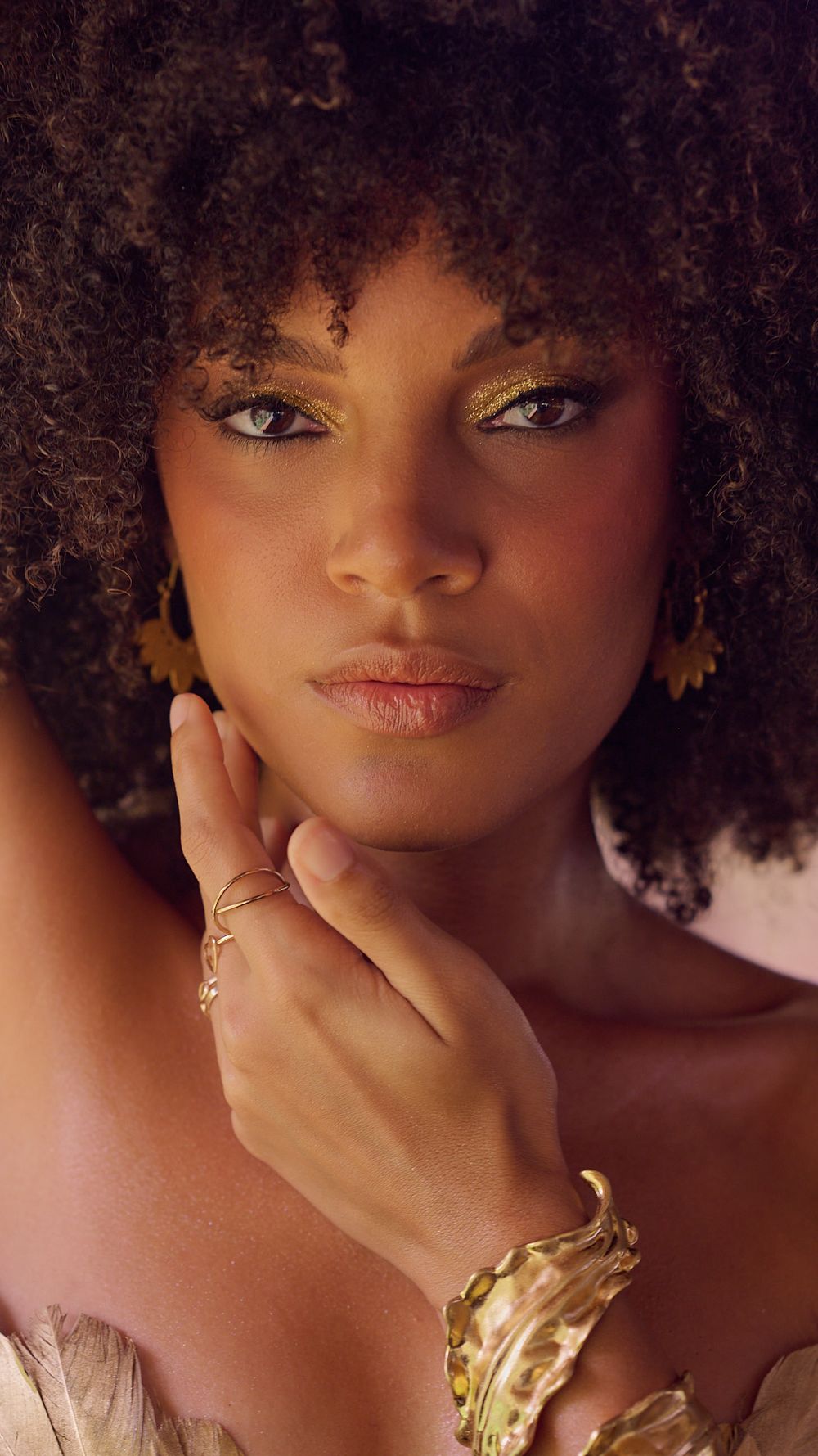 Portrait of a model with natural curly hair wearing gold jewelry and soft makeup against a warm background.