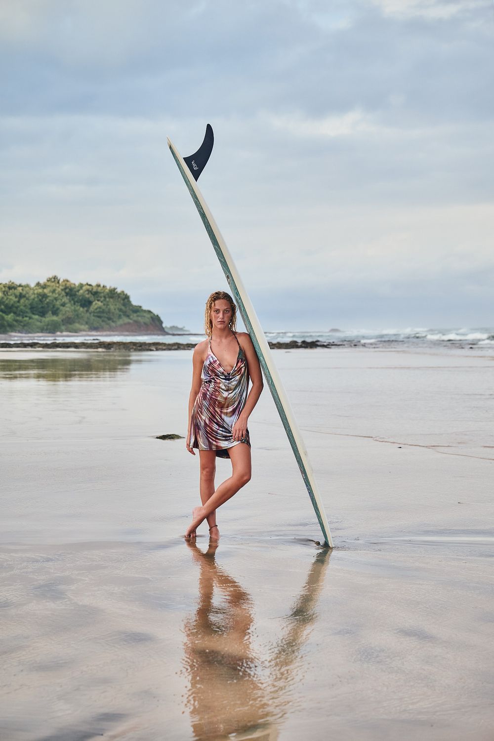 A person stands on a serene beach holding a surfboard while waves roll in during overcast weather.