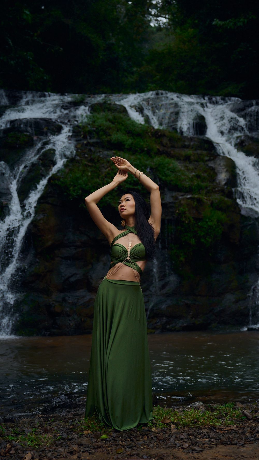 A dancer in an olive green dress performs graceful poses against a scenic waterfall backdrop in a natural setting.