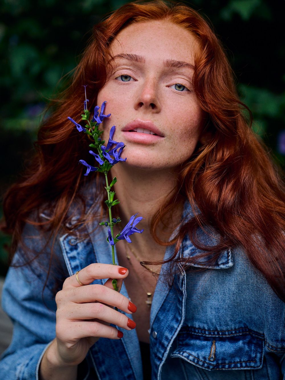 Person with flowing red hair wearing denim jacket holds purple wildflower against blurred green background.