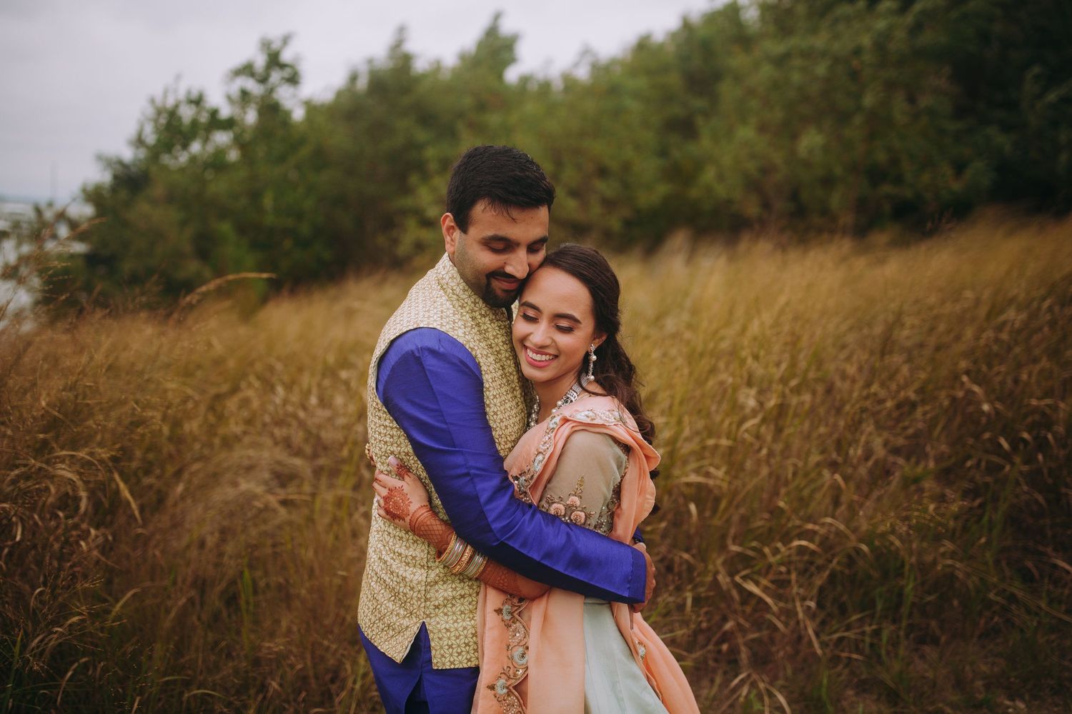 A couple in traditional Indian wedding attire poses together in a scenic autumn setting with golden foliage.