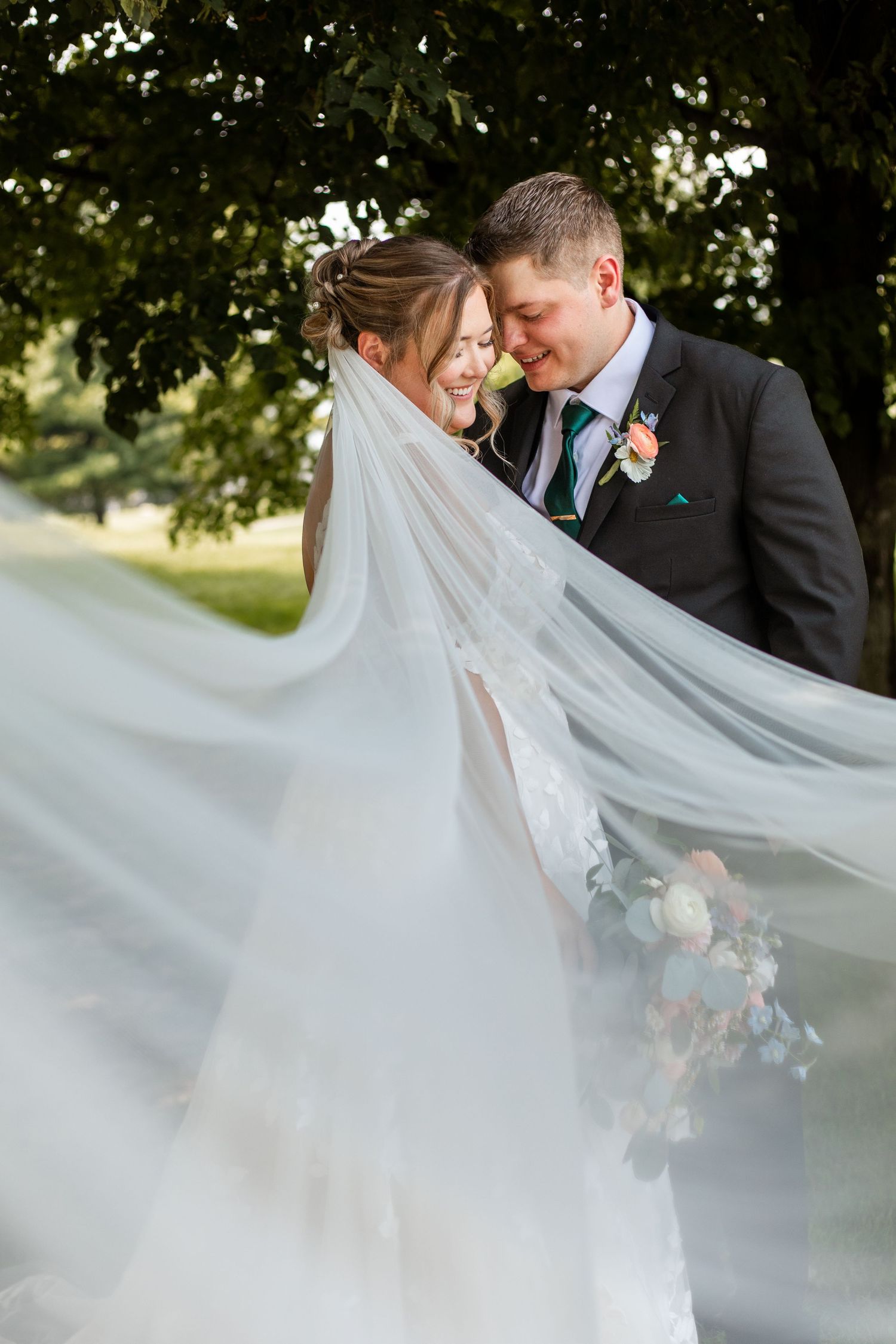 A romantic wedding couple embraces under trees as the bride's flowing veil creates an ethereal effect in the breeze.