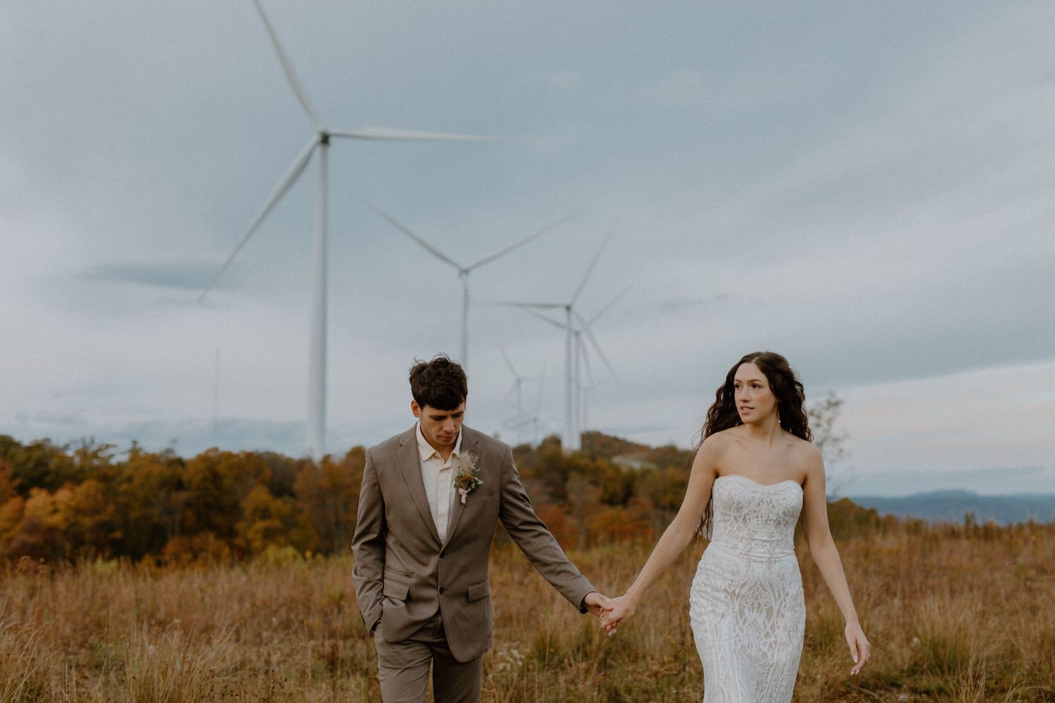 Bride and groom walk hand in hand through an open field with wind turbines and colorful autumn foliage in the background.