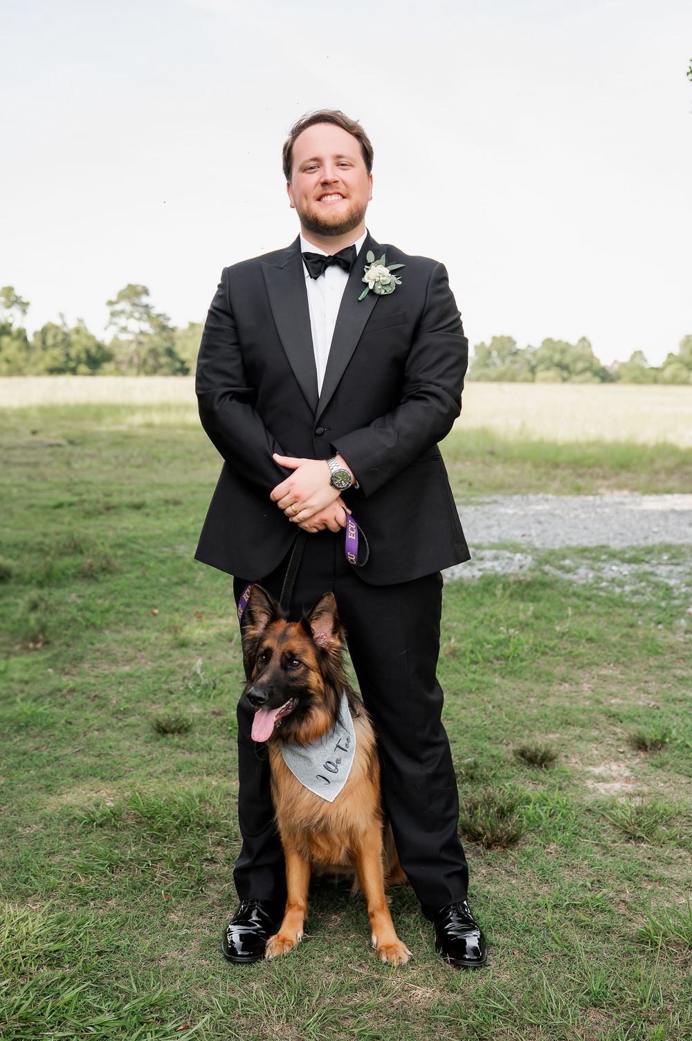 Groom in black tuxedo poses with German Shepherd wearing blue bowtie.