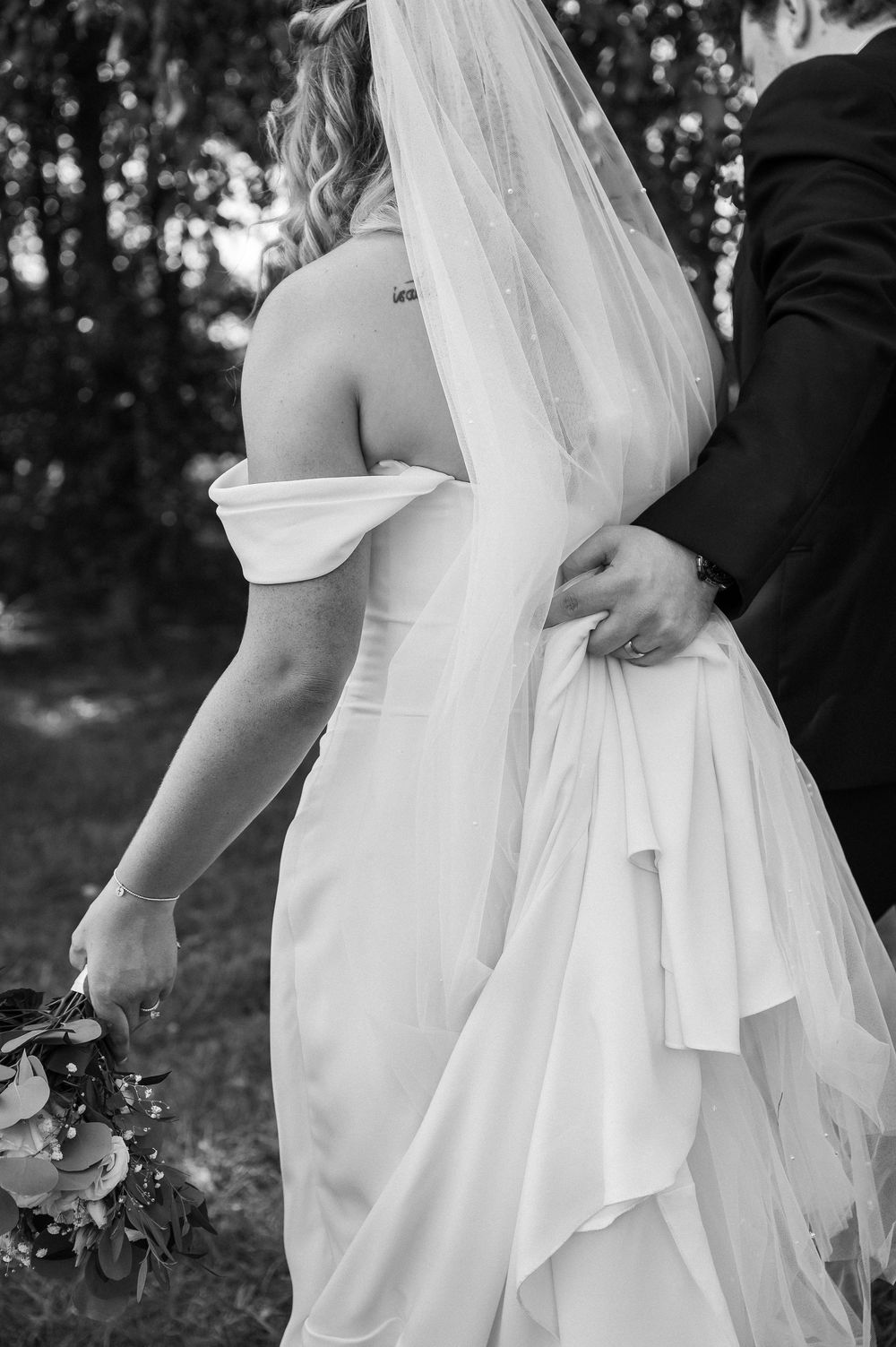 Black and white wedding photo showing the back of a bride's off-shoulder dress and flowing veil in a forest setting.