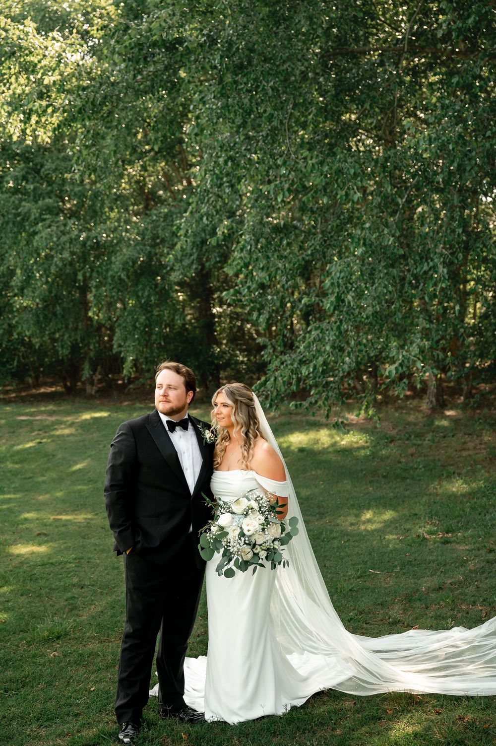 Wedding couple poses in front of lush green trees with bride holding white floral bouquet and wearing long flowing veil.