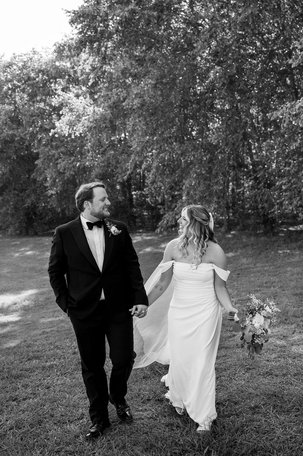 Black and white photo of newlyweds walking hand in hand through a garden setting.