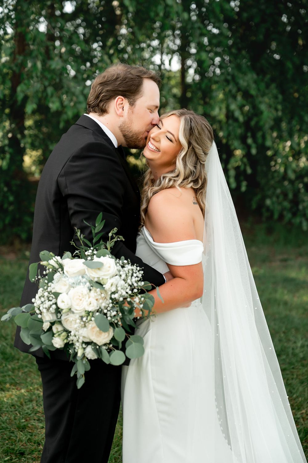 Romantic wedding portrait with couple embracing while holding white and green bouquet in outdoor setting.
