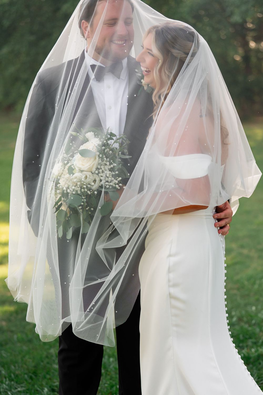 Joyful wedding couple shares a laugh under bride's veil while holding white bouquet.