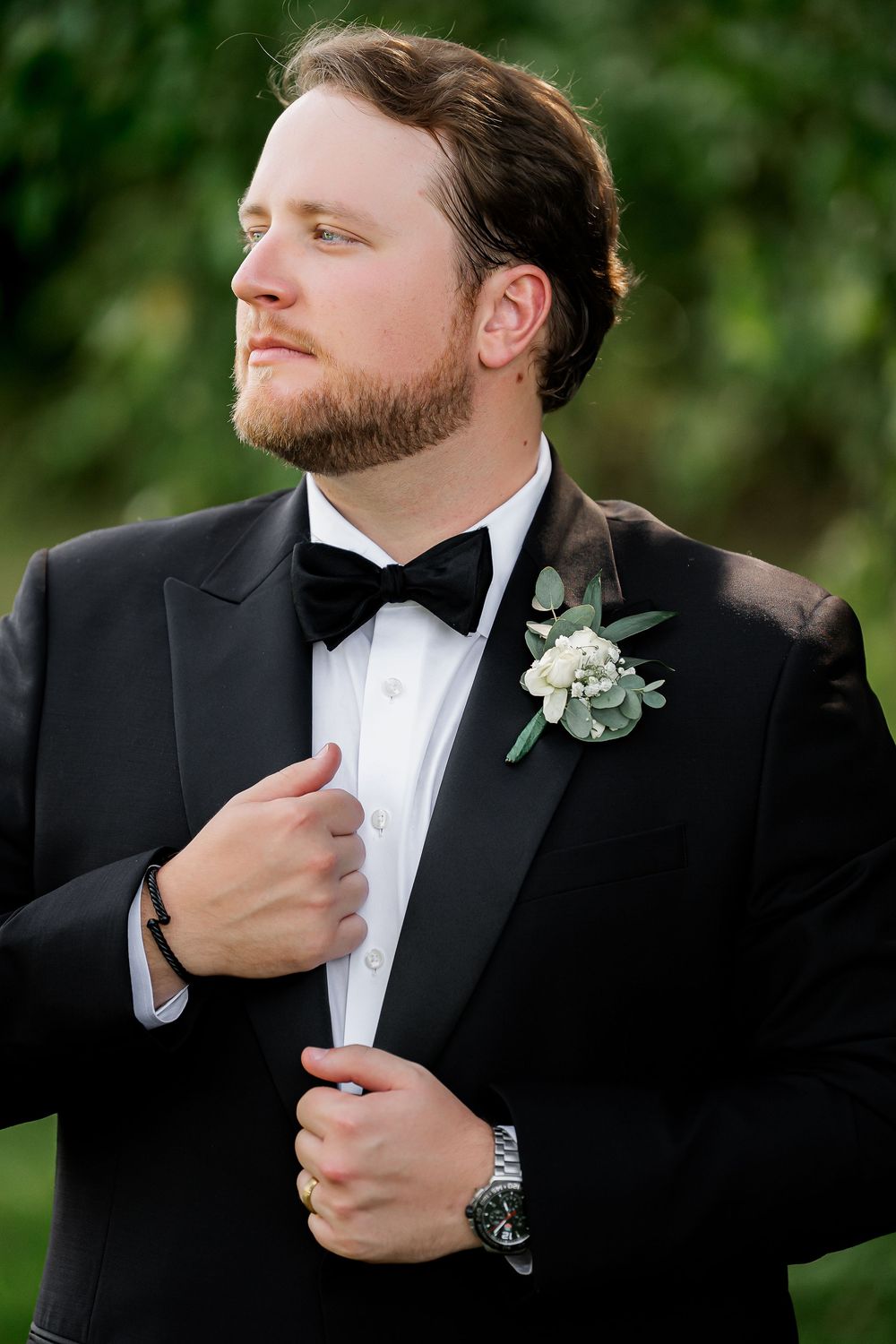 Groom in classic black tuxedo and bowtie adjusts his collar outdoors.