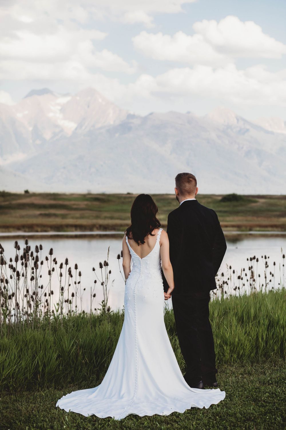 Bride and groom pose near a serene lake surrounded by cattails and mountain vistas.