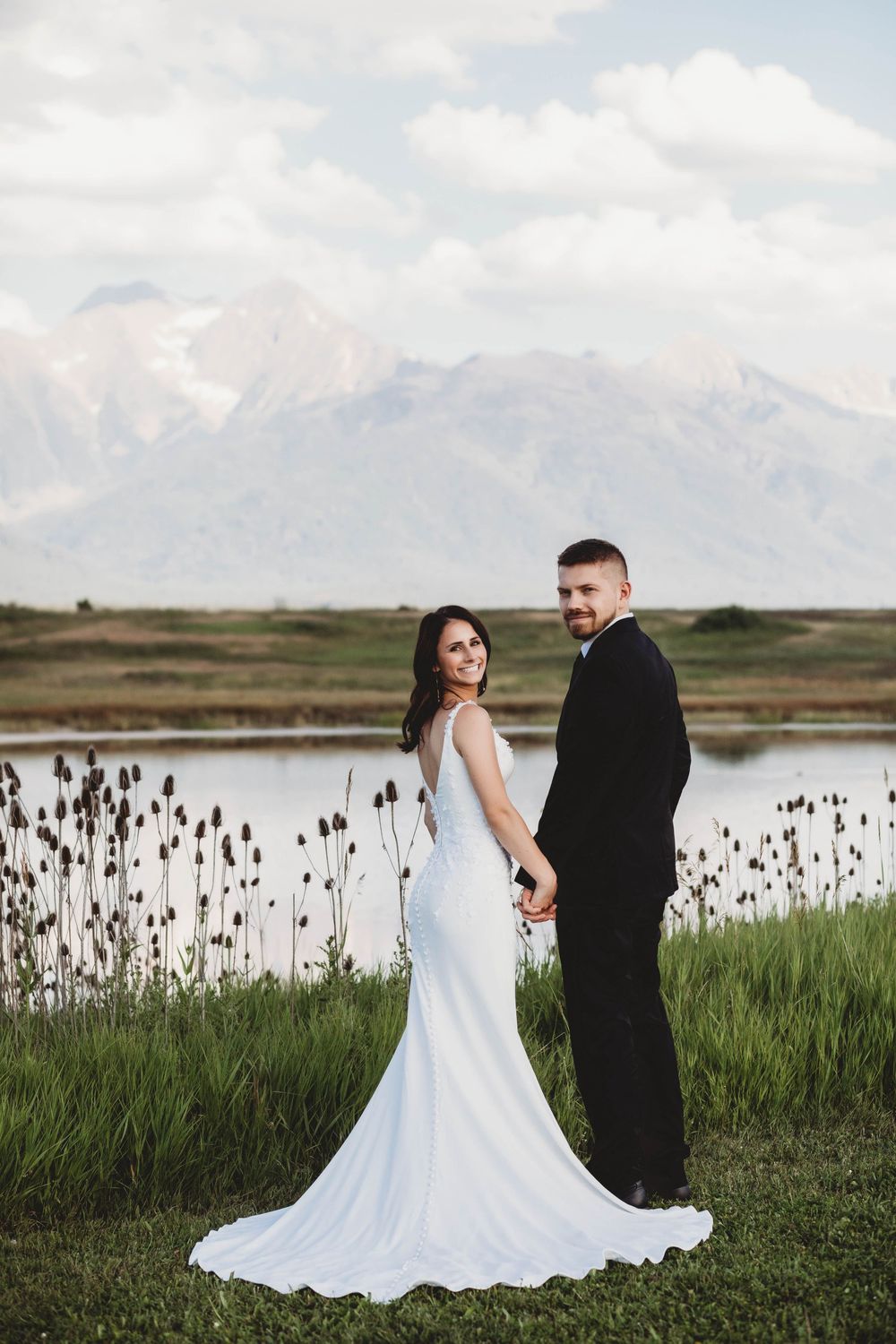 A couple in wedding attire poses against a mountain backdrop with a lake and wildflowers in the foreground.