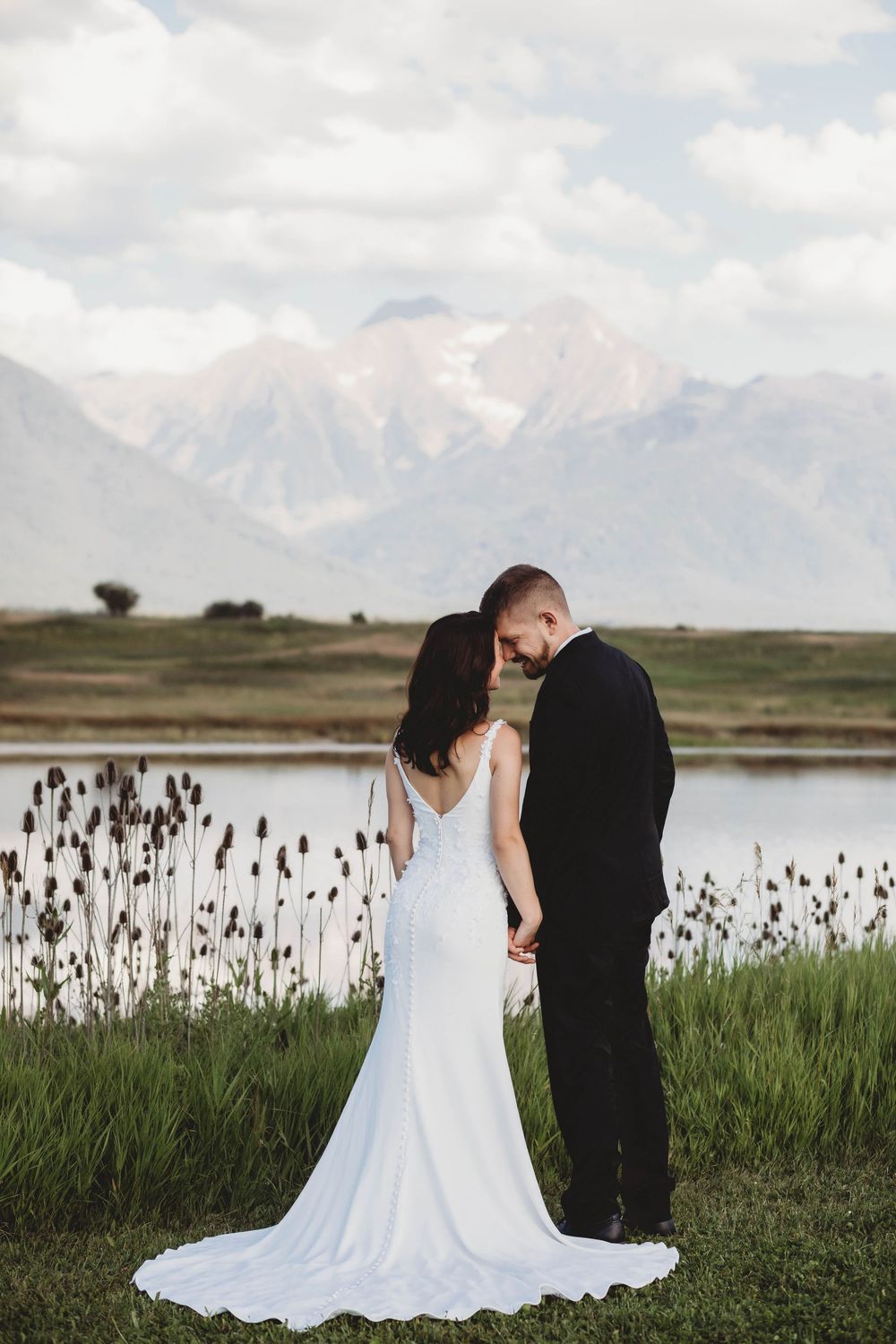 A romantic wedding portrait showing a couple standing by a lake with mountains in the background.