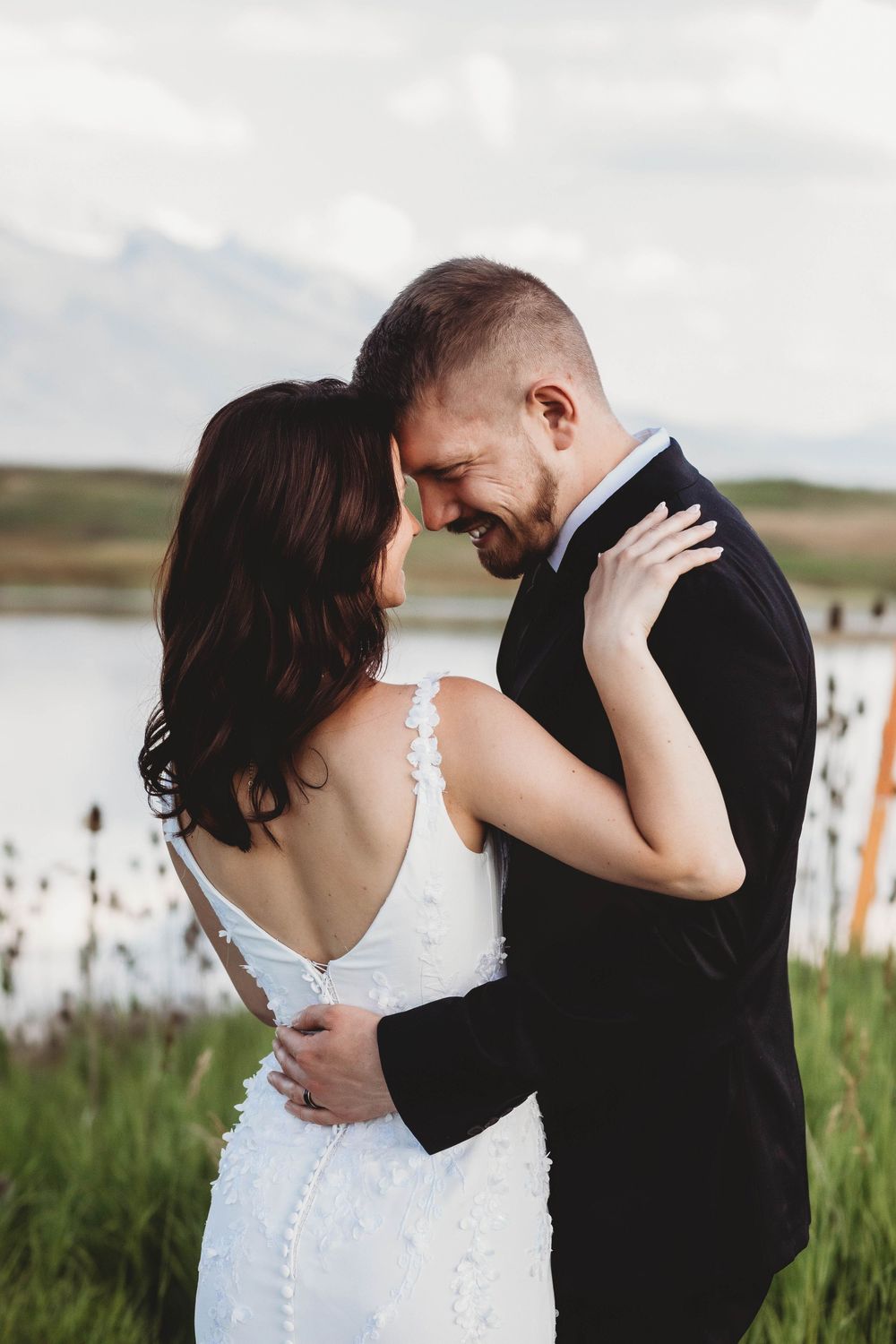 A romantic couple embraces in a field near a lake with cattails and tall grass in the background.