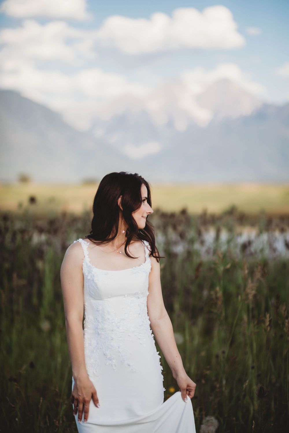 A person in a white dress stands in a field of tall grass with mountains and blue sky in the background.