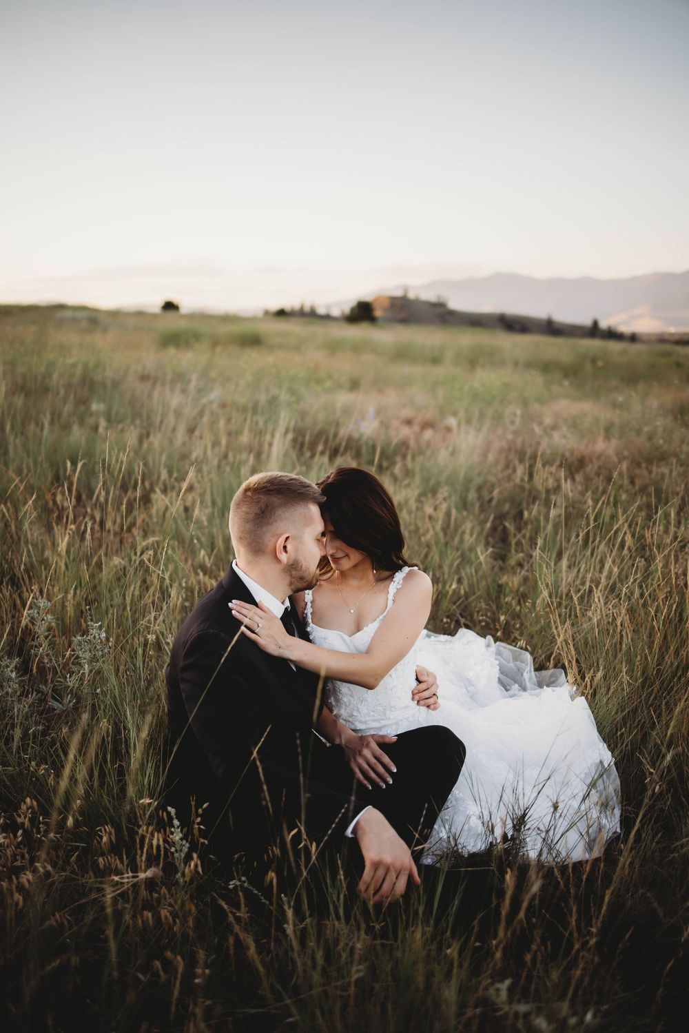 A romantic couple embraces in a field of tall grass at sunset while wearing formal wedding attire.