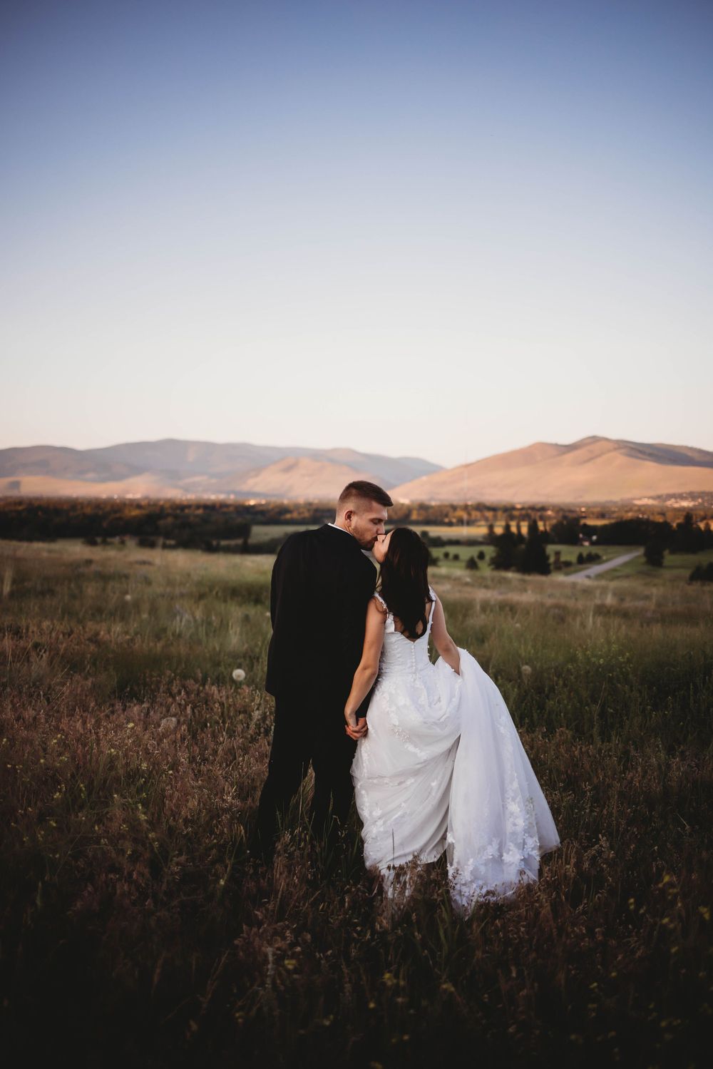 Wedding couple walks through dusky field with mountains silhouetted against evening sky.