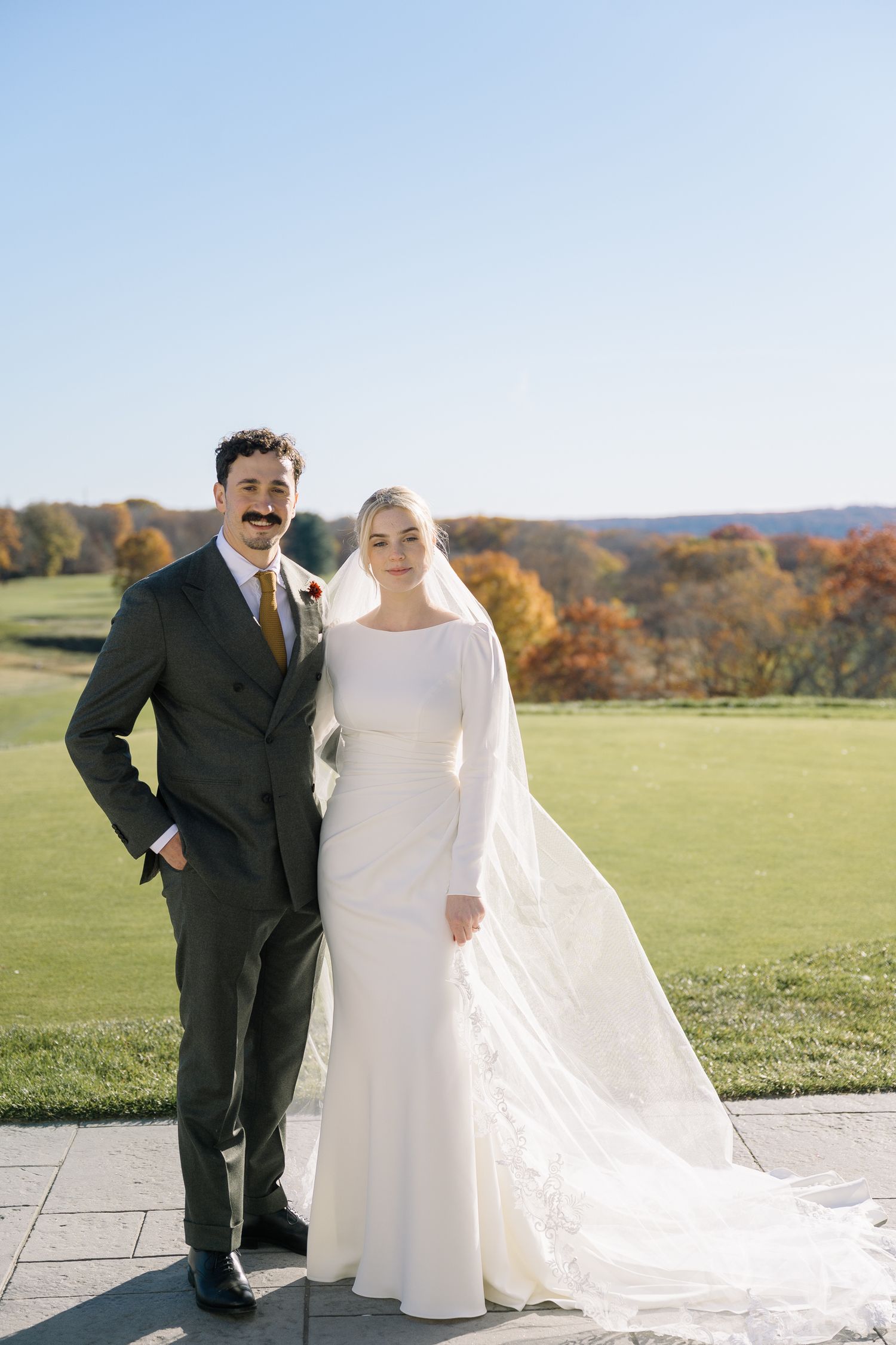 Newlyweds pose together on a sunny autumn day with rolling hills and fall foliage in the background.