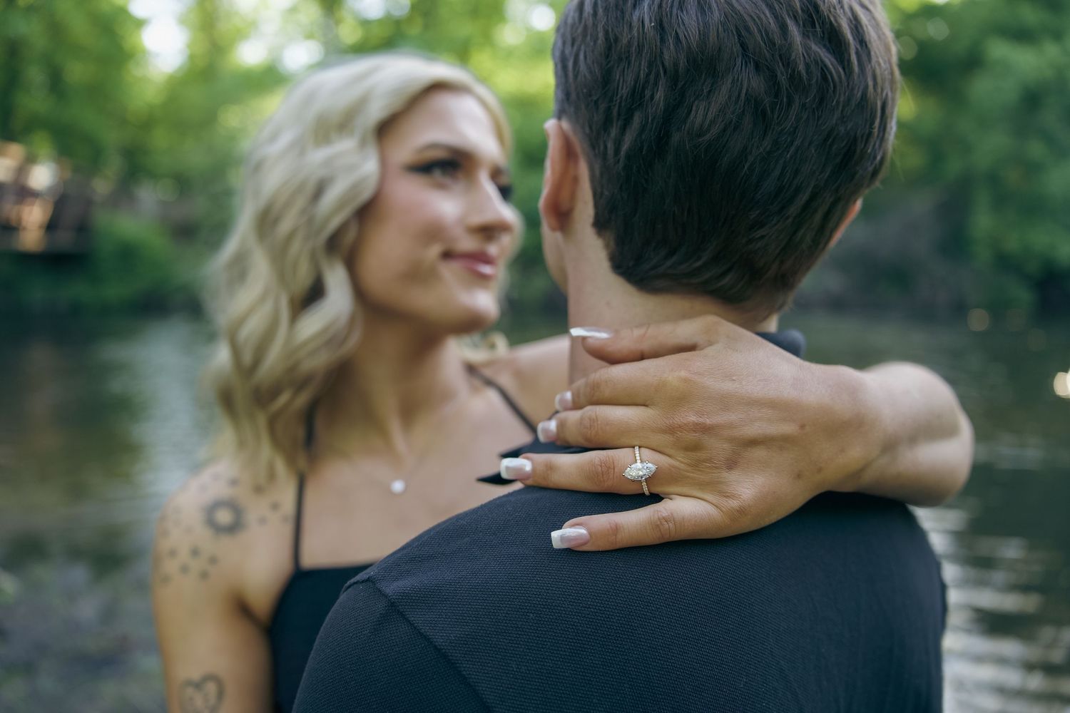 A couple embraces by a lake with a close-up view of an engagement ring on her hand resting on his shoulder.