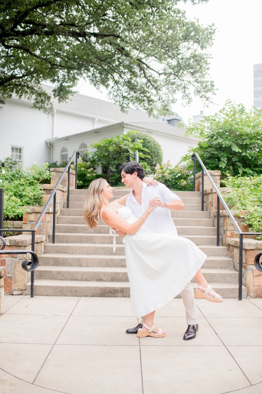 A couple in wedding attire performs a playful dip pose on outdoor concrete steps with greenery in the background.