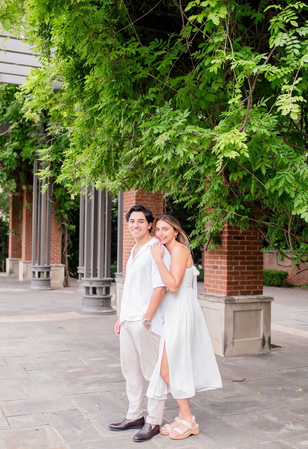 A couple shares a romantic moment together under lush green trees outside a brick building with white trim.