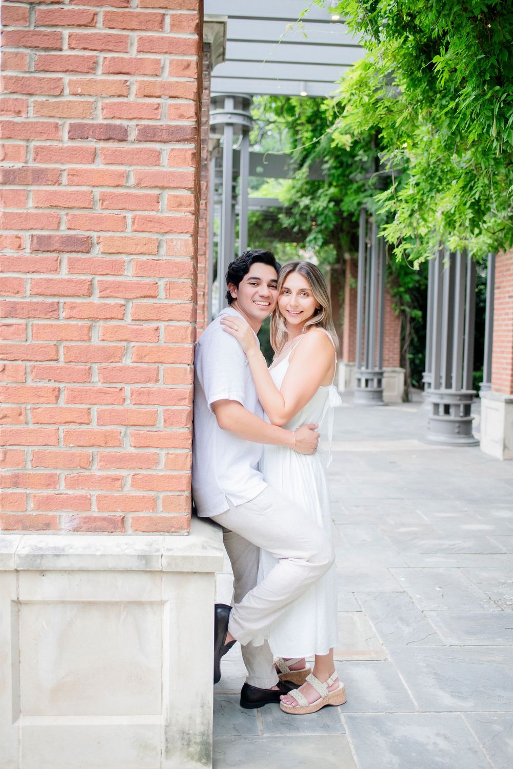 Romantic engagement photo session with couple in white outfits posing against brick wall and columns.
